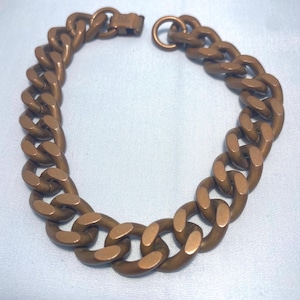 Puede incluir: Un brazalete de cadena de color bronce con cierre de broche. El brazalete presenta eslabones grandes y ovalados. El metal tiene un aspecto ligeramente envejecido. El brazalete se muestra sobre un fondo azul claro.