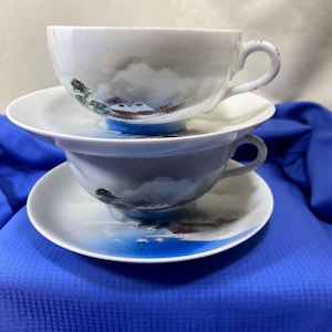 May include: Two white teacups and saucers stacked on top of each other. Each cup and saucer features a landscape design with a blue base, and a handle. The landscape design includes a mountain scene with trees and a blue sky.