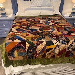May include: A rectangular quilt featuring a patchwork of multi-colored fabric scraps in various textures including velvet, silk, and wool. The quilt is bordered by a wide band of olive green velvet and displays intricate embroidery along the seams.