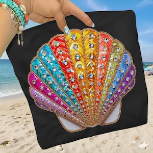 Puede incluir: Toalla de playa negra con un gran diseño de concha marina colorida. La concha está decorada con un arcoíris de colores y brillantes pedrería, delineada con un borde dorado. La toalla se muestra sobre un fondo de playa.