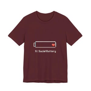 May include: Burgundy t-shirt featuring a pixelated battery graphic with 1% charge and a heart. The text "1% Social Battery" is below the graphic. The shirt has short sleeves and a crew neck.