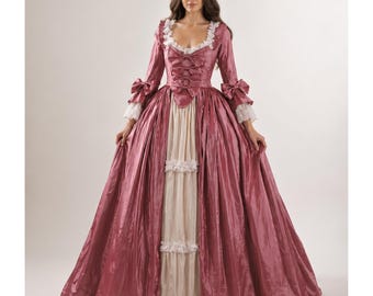 May include: A full-length, antique rose-colored gown with a cream-colored bodice and skirt panel. The dress features long sleeves with bows, lace trim, and a low neckline. The skirt is voluminous, creating a classic, historical silhouette.