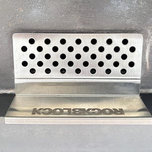 Puede incluir: Un soporte ROCKBLOCK de acero inoxidable con una parte superior perforada y una base con el nombre de la marca grabado. El soporte est&aacute; dise&ntilde;ado para sostener un art&iacute;culo y tiene forma rectangular.