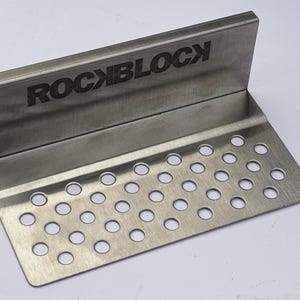 Puede incluir: Una placa met&aacute;lica de acero inoxidable con la palabra "ROCKBLOCK" grabada en el borde superior. La placa tiene un dise&ntilde;o doblado con una secci&oacute;n inferior perforada con numerosos agujeros circulares espaciados.