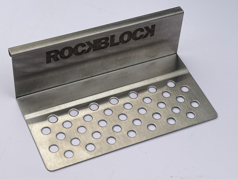 Puede incluir: Una placa met&aacute;lica de acero inoxidable con la palabra "ROCKBLOCK" grabada en el borde superior. La placa tiene un dise&ntilde;o doblado con una secci&oacute;n inferior perforada con numerosos agujeros circulares espaciados.
