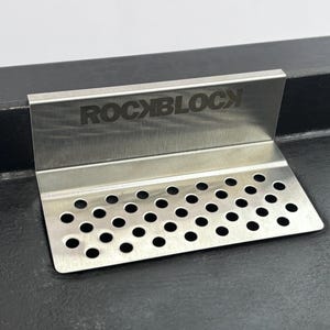 Puede incluir: Un colador de fregadero de cocina de acero inoxidable con el texto "ROCKBLOCK" grabado en la parte superior. El colador tiene una base perforada con numerosos agujeros peque&ntilde;os y espaciados uniformemente. El art&iacute;culo est&aacute; dise&ntilde;ado para colocarse en un &aacute;ngulo de 90 grados.