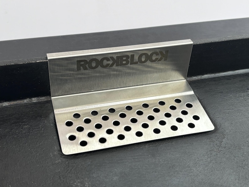 Puede incluir: Un colador de fregadero de cocina de acero inoxidable con el texto "ROCKBLOCK" grabado en la parte superior. El colador tiene una base perforada con numerosos agujeros peque&ntilde;os y espaciados uniformemente. El art&iacute;culo est&aacute; dise&ntilde;ado para colocarse en un &aacute;ngulo de 90 grados.