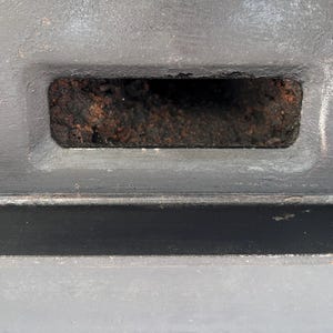 Puede incluir: Primer plano de una abertura rectangular en una superficie gris. La abertura est&aacute; llena de un material oscuro y texturizado. La superficie circundante tambi&eacute;n es gris, con sutiles variaciones de tono y textura.
