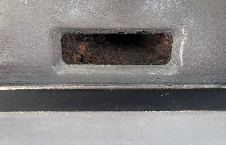 Puede incluir: Primer plano de una abertura rectangular en una superficie gris. La abertura est&aacute; llena de un material oscuro y texturizado. La superficie circundante tambi&eacute;n es gris, con sutiles variaciones de tono y textura.
