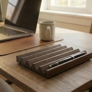 May include: A wooden desk organizer with a pen in it sits on a wooden desk. A laptop, a mug, and a small potted succulent are also on the desk. The organizer has a series of angled slots for holding pens or pencils. The desk is in a well-lit room.