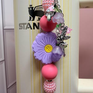 May include: A decorative tumbler charm featuring a large purple flower bead, pink and textured light pink spherical beads, and silver-toned metal charms including a fairy, small mushrooms, and translucent purple bell-shaped flower pendants.