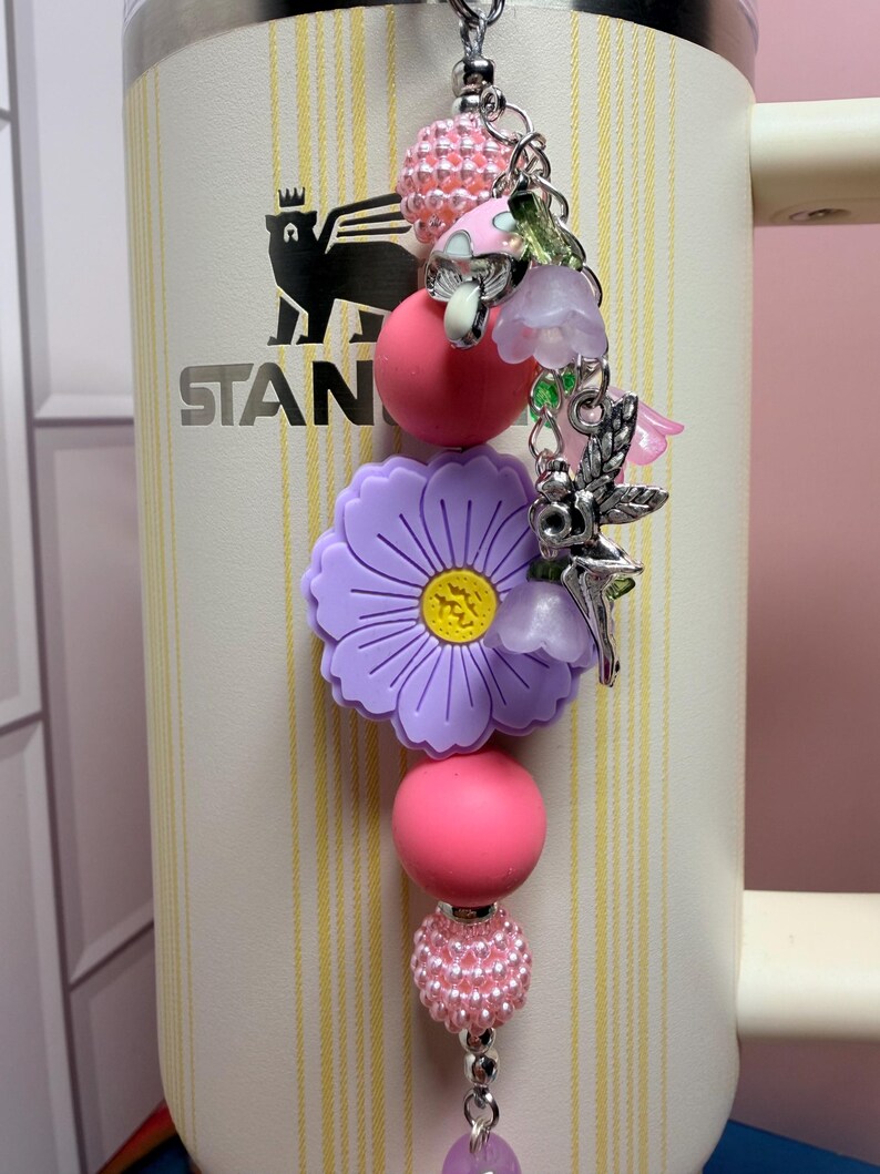 May include: A decorative tumbler charm featuring a large purple flower bead, pink and textured light pink spherical beads, and silver-toned metal charms including a fairy, small mushrooms, and translucent purple bell-shaped flower pendants.