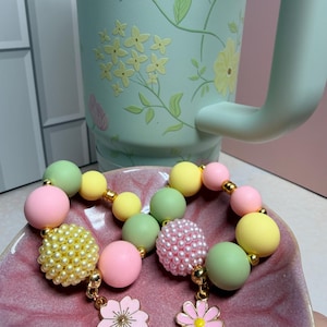May include: A pastel-colored beaded bracelet with gold accents and flower charms. The bracelet features pink, yellow, and green beads. A light green floral-patterned mug is in the background, and the bracelet is displayed on a pink dish.