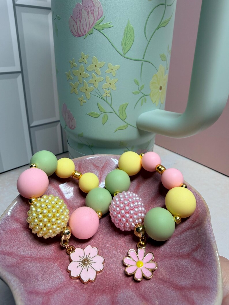 May include: A pastel-colored beaded bracelet with gold accents and flower charms. The bracelet features pink, yellow, and green beads. A light green floral-patterned mug is in the background, and the bracelet is displayed on a pink dish.