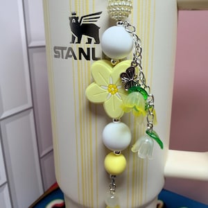 May include: A decorative tumbler charm featuring a silver-tone metal chain with a white pearl-textured bead, two smooth white silicone beads, a yellow silicone flower, a small silver butterfly charm, and two translucent yellow bell-shaped flower beads.