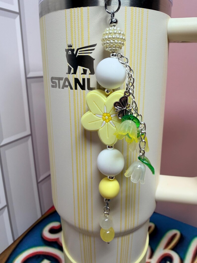 May include: A decorative tumbler charm featuring a silver-tone metal chain with a white pearl-textured bead, two smooth white silicone beads, a yellow silicone flower, a small silver butterfly charm, and two translucent yellow bell-shaped flower beads.