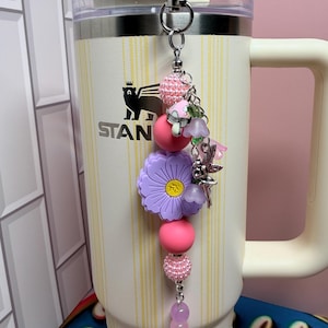 May include: A decorative tumbler charm featuring a silver metal clip, pink textured beads, a large purple flower-shaped bead with a yellow center, and smaller charms including a mushroom, a fairy, and light purple translucent beads.