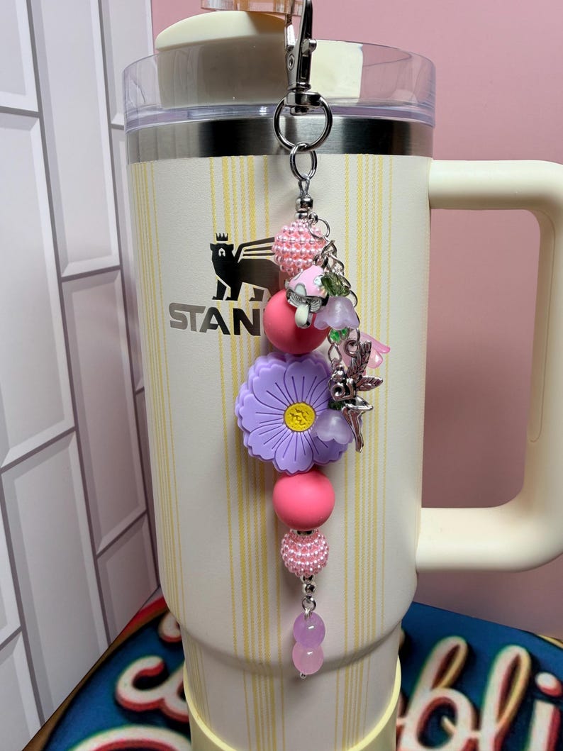 May include: A decorative tumbler charm featuring a silver metal clip, pink textured beads, a large purple flower-shaped bead with a yellow center, and smaller charms including a mushroom, a fairy, and light purple translucent beads.