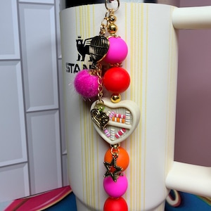 K&ouml;nnte beinhalten: Ein dekorativer Anh&auml;nger mit rotem Clip, goldener Kette und bunten Perlen. Der Anh&auml;nger hat einen rosa Pompon, einen goldenen Vogelk&auml;fig, ein wei&szlig;es Herz mit bunten Details und einen goldenen Stern. Der Anh&auml;nger ist an einem wei&szlig;en Becher befestigt.