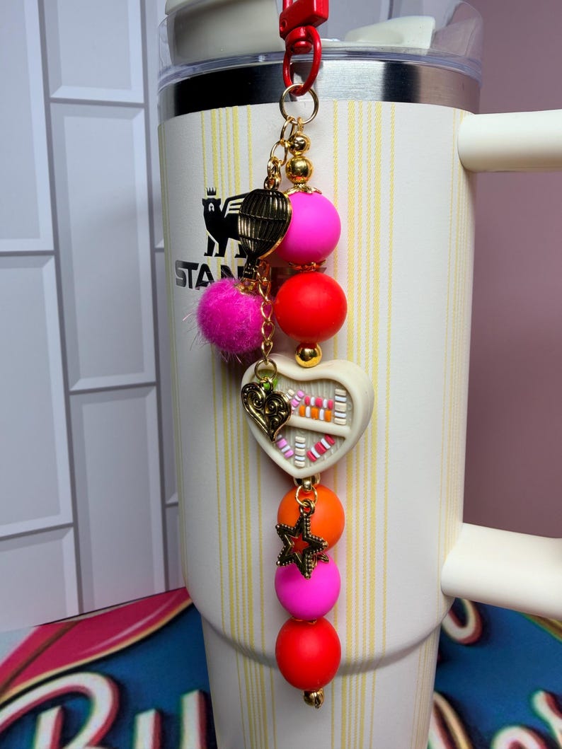 K&ouml;nnte beinhalten: Ein dekorativer Anh&auml;nger mit rotem Clip, goldener Kette und bunten Perlen. Der Anh&auml;nger hat einen rosa Pompon, einen goldenen Vogelk&auml;fig, ein wei&szlig;es Herz mit bunten Details und einen goldenen Stern. Der Anh&auml;nger ist an einem wei&szlig;en Becher befestigt.