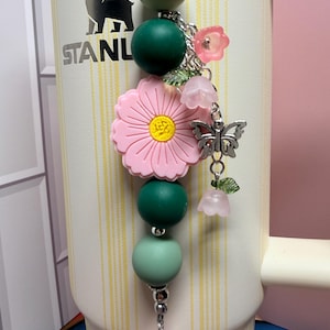 May include: A decorative tumbler charm featuring a vertical arrangement of matte sage and dark green silicone beads, a large pink flower bead with a yellow center, and silver-toned metal charms including a butterfly and small translucent pink flowers.