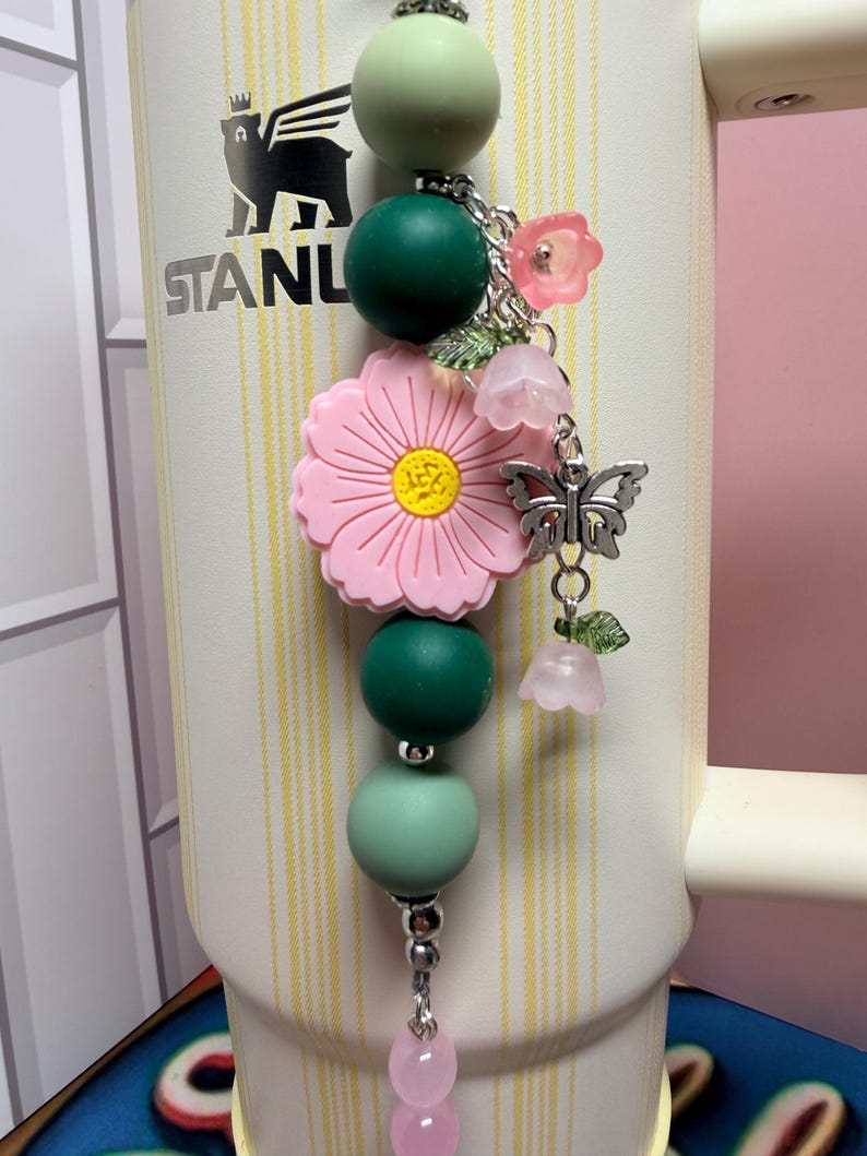 May include: A decorative tumbler charm featuring a vertical arrangement of matte sage and dark green silicone beads, a large pink flower bead with a yellow center, and silver-toned metal charms including a butterfly and small translucent pink flowers.