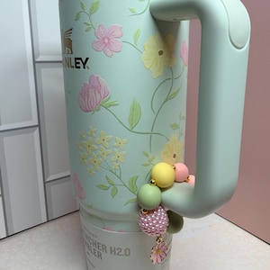 May include: A mint green Stanley Quencher H2.0 Flowstate tumbler with a floral design in pink and yellow. The tumbler has a handle and a decorative beaded charm with a flower. The top of the tumbler has a gold band.