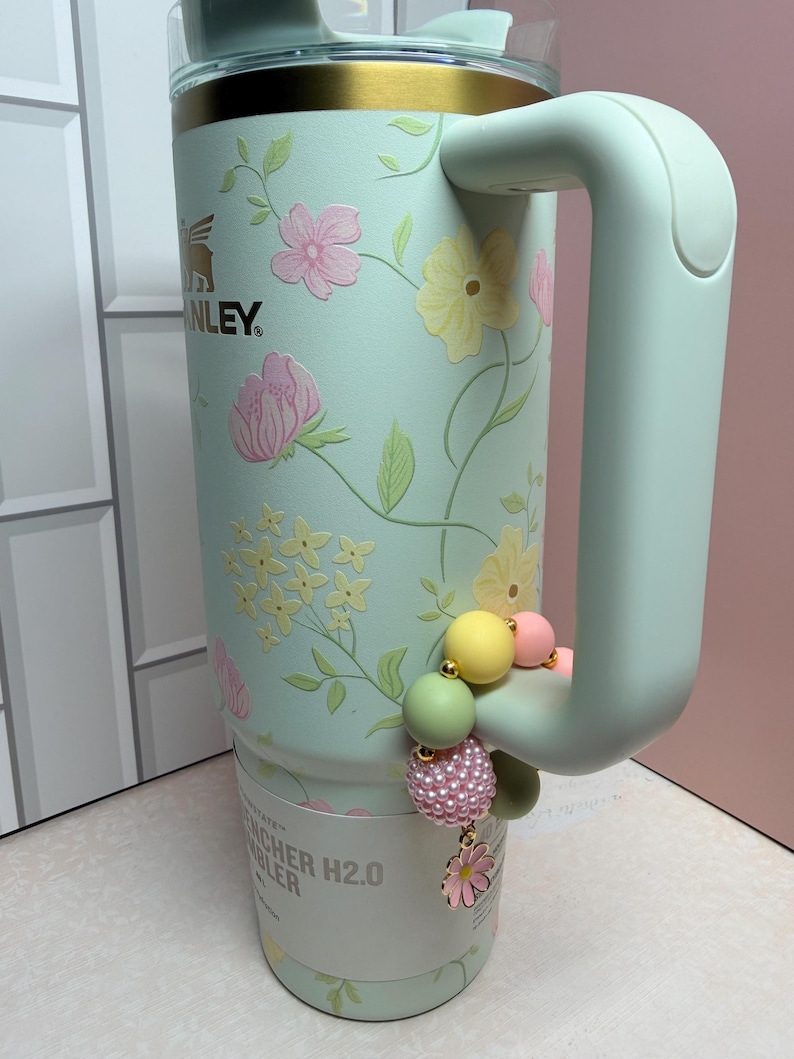 May include: A mint green Stanley Quencher H2.0 Flowstate tumbler with a floral design in pink and yellow. The tumbler has a handle and a decorative beaded charm with a flower. The top of the tumbler has a gold band.