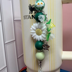 May include: A decorative tumbler charm featuring a white daisy, green and light green silicone beads, a floral patterned bead, and translucent bell-shaped flower charms. A small silver butterfly charm hangs from a silver chain, attached to a cream-colored Stanley tumbler with yellow vertical stripes and a black bear logo.