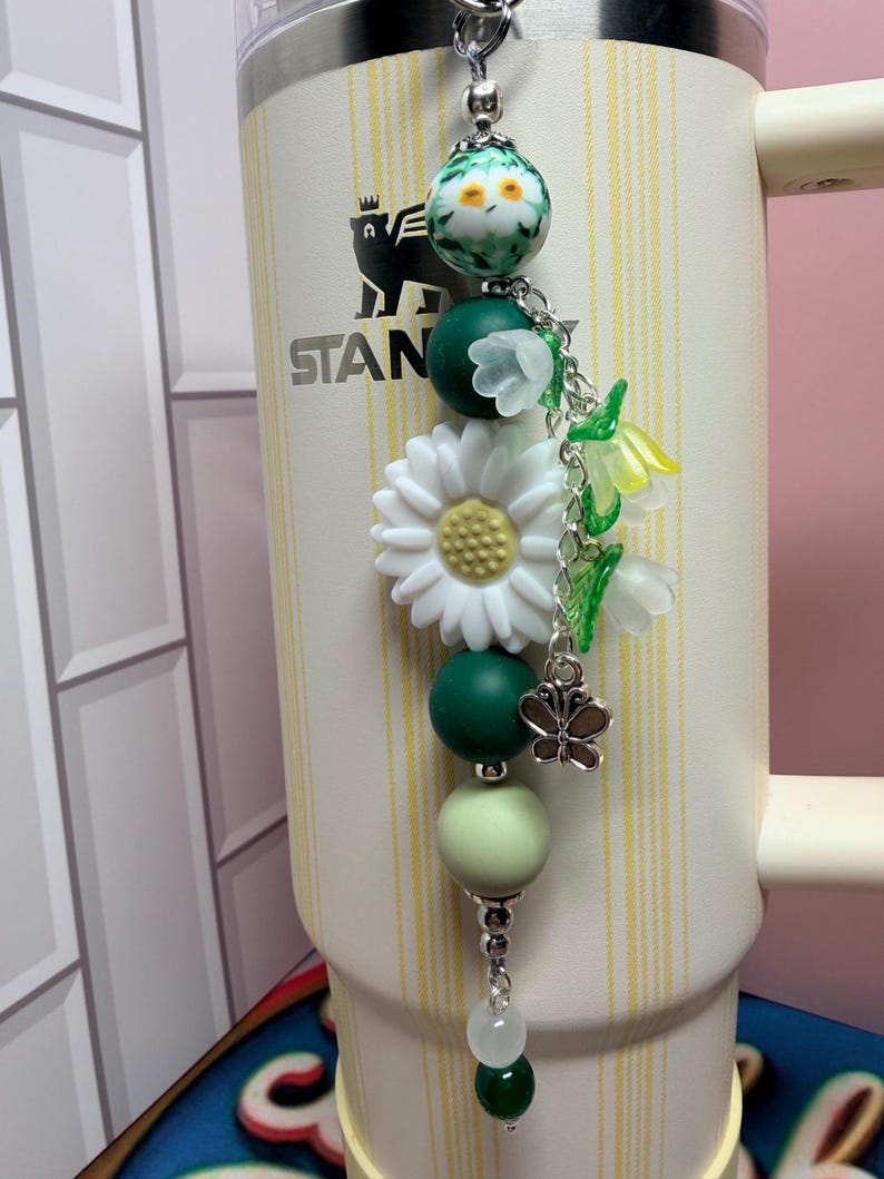 May include: A decorative tumbler charm featuring a white daisy, green and light green silicone beads, a floral patterned bead, and translucent bell-shaped flower charms. A small silver butterfly charm hangs from a silver chain, attached to a cream-colored Stanley tumbler with yellow vertical stripes and a black bear logo.