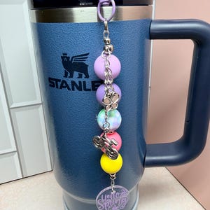 May include: A navy blue Stanley travel mug with a purple clip and a beaded charm. The charm includes a bicycle charm, colourful beads, and a clear tag with the words "Hello Spring".