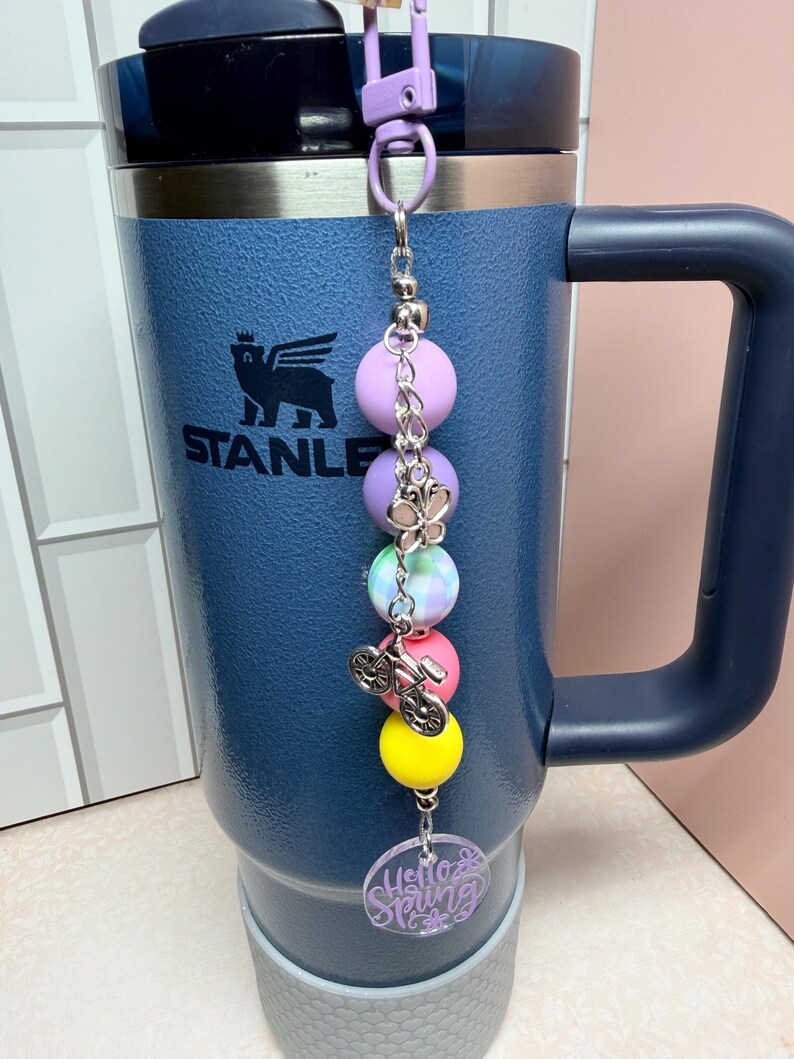 May include: A navy blue Stanley travel mug with a purple clip and a beaded charm. The charm includes a bicycle charm, colourful beads, and a clear tag with the words "Hello Spring".