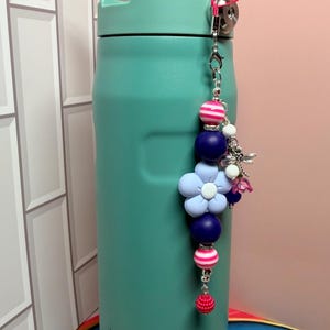 May include: A teal Stanley water bottle with a dark blue handle, featuring a decorative beaded charm attached to the top. The charm includes pink and white striped beads, solid dark blue spheres, a light blue flower bead, and a silver dragonfly.