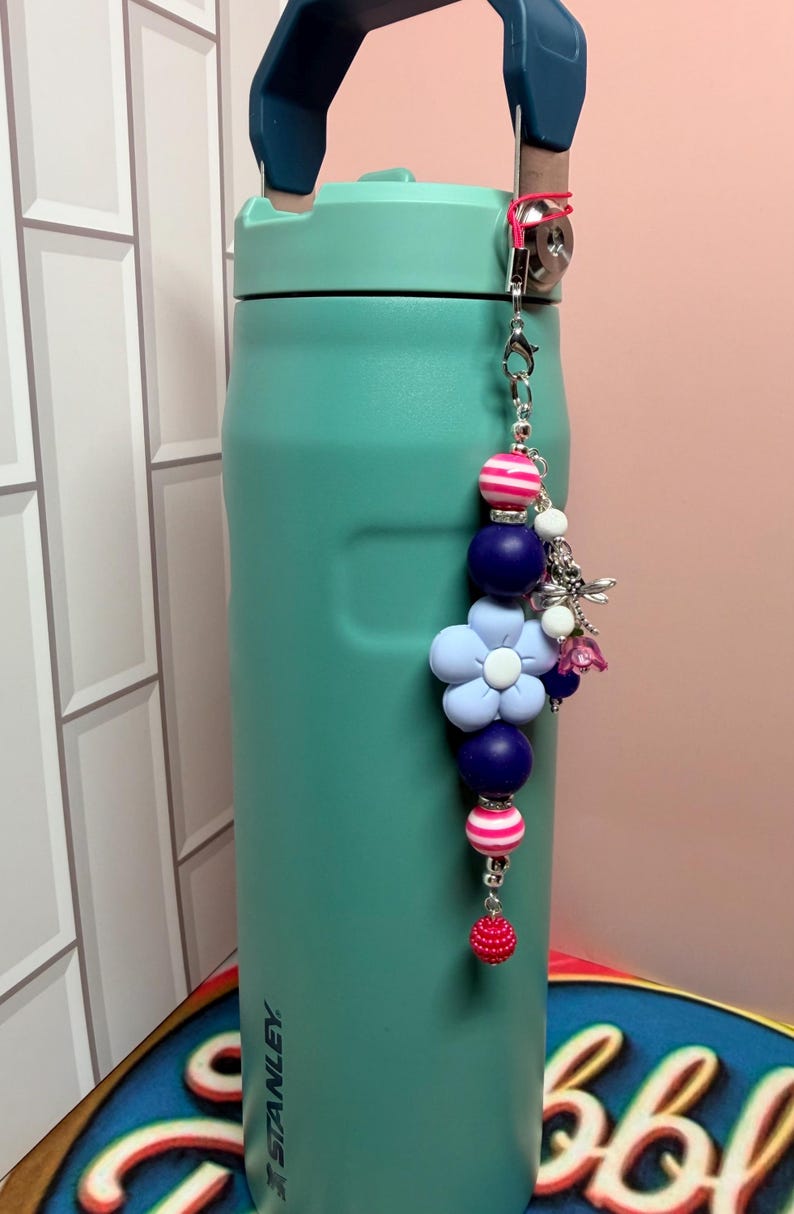 May include: A teal Stanley water bottle with a dark blue handle, featuring a decorative beaded charm attached to the top. The charm includes pink and white striped beads, solid dark blue spheres, a light blue flower bead, and a silver dragonfly.