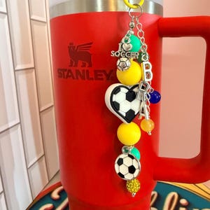 Puede incluir: Un vaso rojo Stanley con asa tiene un dije de f&uacute;tbol sujeto a un clip amarillo. El dije incluye cuentas amarillas, verde azulado y azules, un dije de coraz&oacute;n con forma de bal&oacute;n de f&uacute;tbol, un bal&oacute;n redondo y cuentas con las letras plateadas BRA.