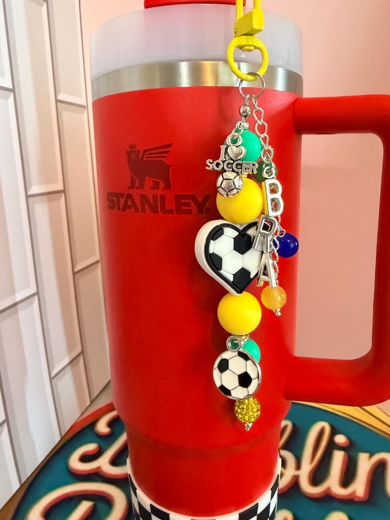 Puede incluir: Un vaso rojo Stanley con asa tiene un dije de f&uacute;tbol sujeto a un clip amarillo. El dije incluye cuentas amarillas, verde azulado y azules, un dije de coraz&oacute;n con forma de bal&oacute;n de f&uacute;tbol, un bal&oacute;n redondo y cuentas con las letras plateadas BRA.