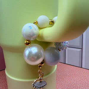 Op de afbeelding: Een witte armband met kralen en gouden accenten, met een ballerina-bedel. De armband heeft grote, ronde, parelachtige kralen en kleinere witte kralen. De bedel is een gouden ballerina met een witte tutu en strass-accenten.