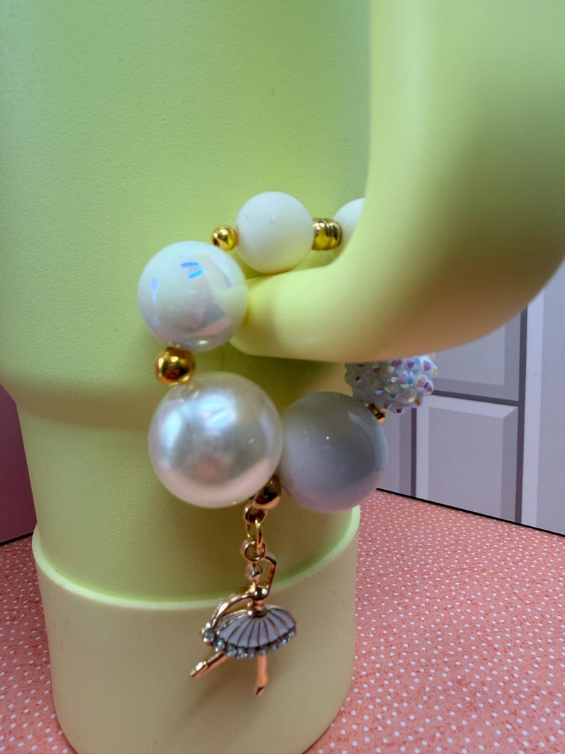 Op de afbeelding: Een witte armband met kralen en gouden accenten, met een ballerina-bedel. De armband heeft grote, ronde, parelachtige kralen en kleinere witte kralen. De bedel is een gouden ballerina met een witte tutu en strass-accenten.