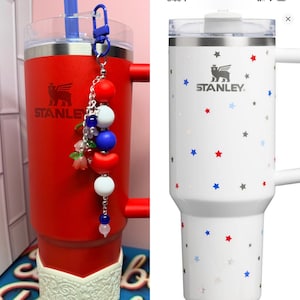 Op de afbeelding: Twee Stanley-bekers. E&eacute;n is rood met een witte basis en een kralenbedel. De andere is wit met een sterrenpatroon. Beide hebben het Stanley-logo en een handvat. De rode beker heeft een blauw rietje.