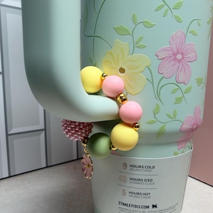 May include: A light green floral-patterned tumbler with a beaded charm bracelet. The bracelet features yellow, pink, and green beads, gold accents, and a flower charm. The tumbler has a handle and a white label with text.
