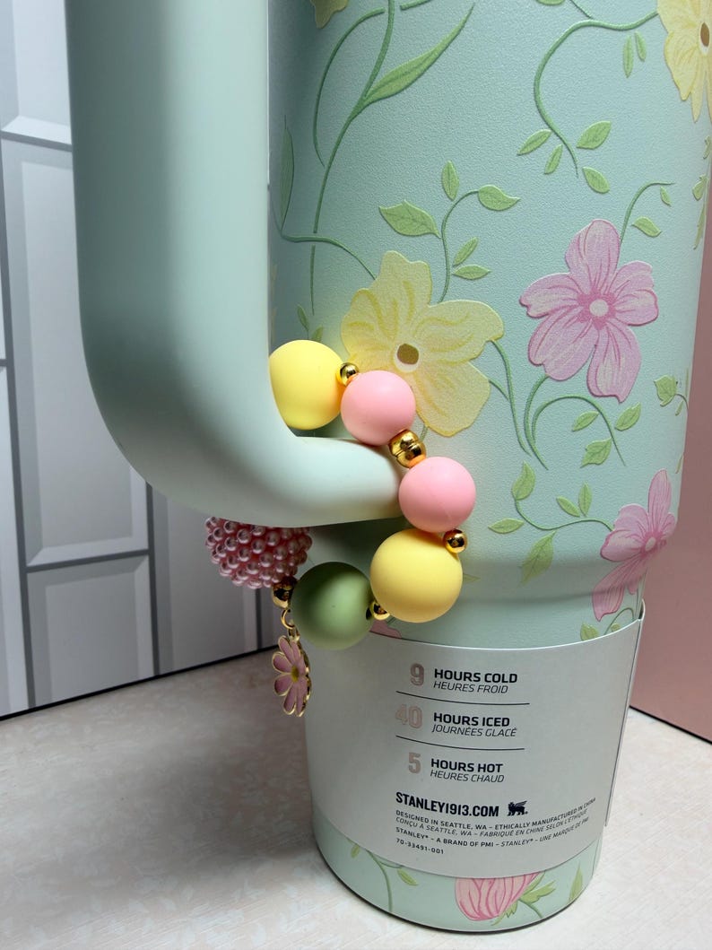 May include: A light green floral-patterned tumbler with a beaded charm bracelet. The bracelet features yellow, pink, and green beads, gold accents, and a flower charm. The tumbler has a handle and a white label with text.