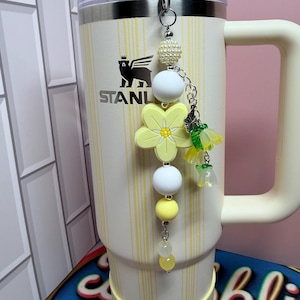 May include: A cream-colored tumbler with a handle and vertical yellow stripes, featuring a logo with a bear and the text STANLEY. A beaded charm hangs from the lid, showcasing a large yellow flower, white and yellow beads, and small floral charms.