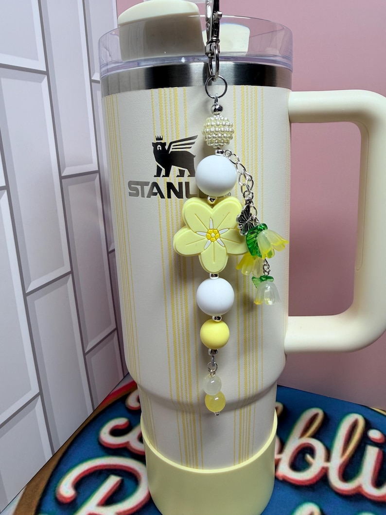 May include: A cream-colored tumbler with a handle and vertical yellow stripes, featuring a logo with a bear and the text STANLEY. A beaded charm hangs from the lid, showcasing a large yellow flower, white and yellow beads, and small floral charms.