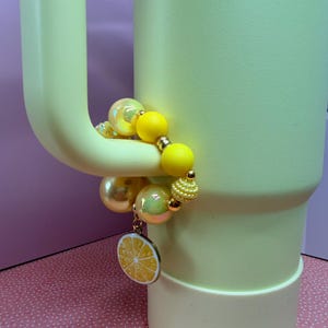 Op de afbeelding: Een lichtgele beker met bijpassende handgreep, versierd met een kralenarmband. De armband heeft gele, gouden en iriserende kralen, samen met een bedel in de vorm van een citroenschijf. De beker staat op een roze ondergrond.