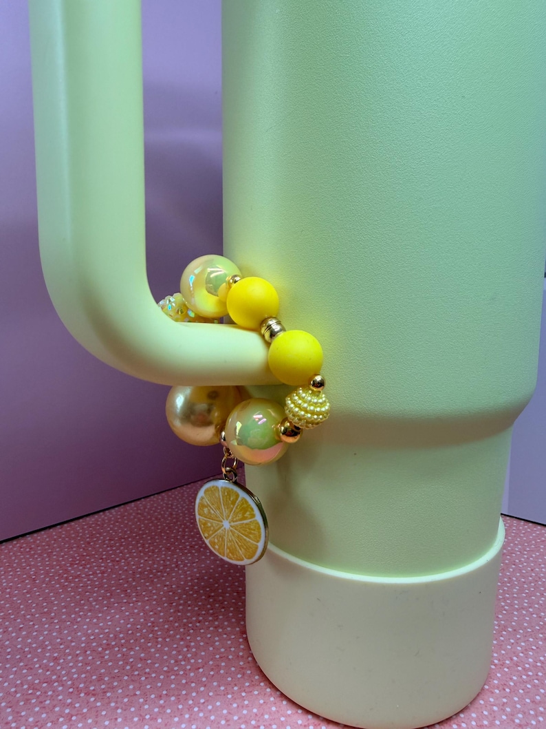 Op de afbeelding: Een lichtgele beker met bijpassende handgreep, versierd met een kralenarmband. De armband heeft gele, gouden en iriserende kralen, samen met een bedel in de vorm van een citroenschijf. De beker staat op een roze ondergrond.