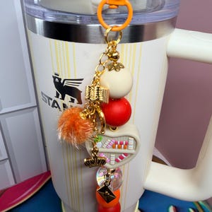 K&ouml;nnte beinhalten: Ein cremefarbener Stanley-Becher mit einem dekorativen Anh&auml;nger. Der Anh&auml;nger hat eine goldene Kette mit einem wei&szlig;en Ball, einem roten Ball, einem orangefarbenen Pompon und einem kleinen B&uuml;cherregal-Design. Der Anh&auml;nger ist an einem orangefarbenen Karabiner befestigt.