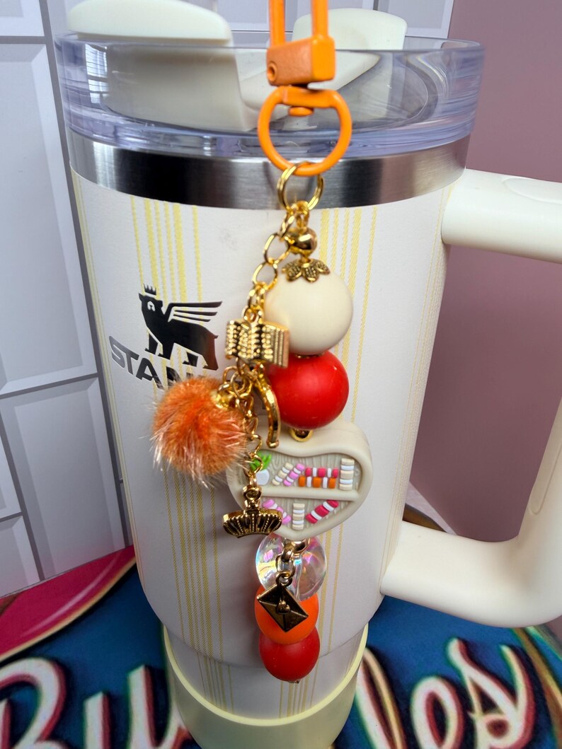 K&ouml;nnte beinhalten: Ein cremefarbener Stanley-Becher mit einem dekorativen Anh&auml;nger. Der Anh&auml;nger hat eine goldene Kette mit einem wei&szlig;en Ball, einem roten Ball, einem orangefarbenen Pompon und einem kleinen B&uuml;cherregal-Design. Der Anh&auml;nger ist an einem orangefarbenen Karabiner befestigt.