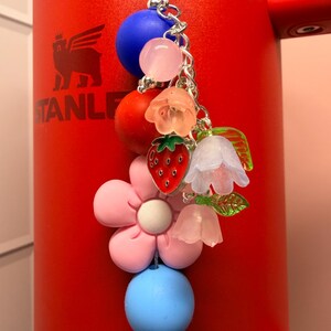 Op de afbeelding: Een kleurrijke bedel met diverse kralen en bedels, waaronder een rode aardbei, een roze bloem en blauwe en roze kralen. De bedel is bevestigd aan een zilveren ketting en hangt voor een rode beker.