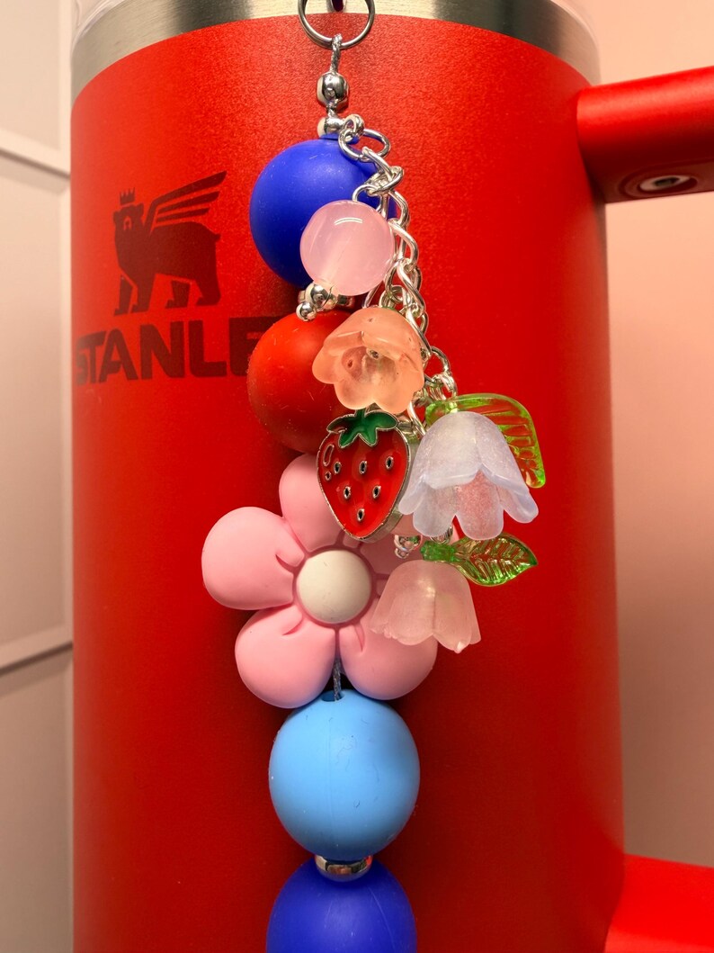 Op de afbeelding: Een kleurrijke bedel met diverse kralen en bedels, waaronder een rode aardbei, een roze bloem en blauwe en roze kralen. De bedel is bevestigd aan een zilveren ketting en hangt voor een rode beker.