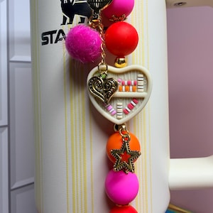 K&ouml;nnte beinhalten: Ein dekorativer Charm mit einer goldenen Kette, einem rosa Pompon und bunten Perlen. Der Charm hat einen herzf&ouml;rmigen Anh&auml;nger mit einem B&uuml;cherregal-Design und einem goldenen Stern. Der Charm ist an einem wei&szlig;en und beigen Stanley-Becher befestigt.